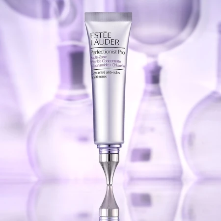 Estee Lauder Perfectionist Pro Multi-Zone Wrinkle Concentrate Niacinamide + Chlorella tube on a reflective surface with blurred flasks.