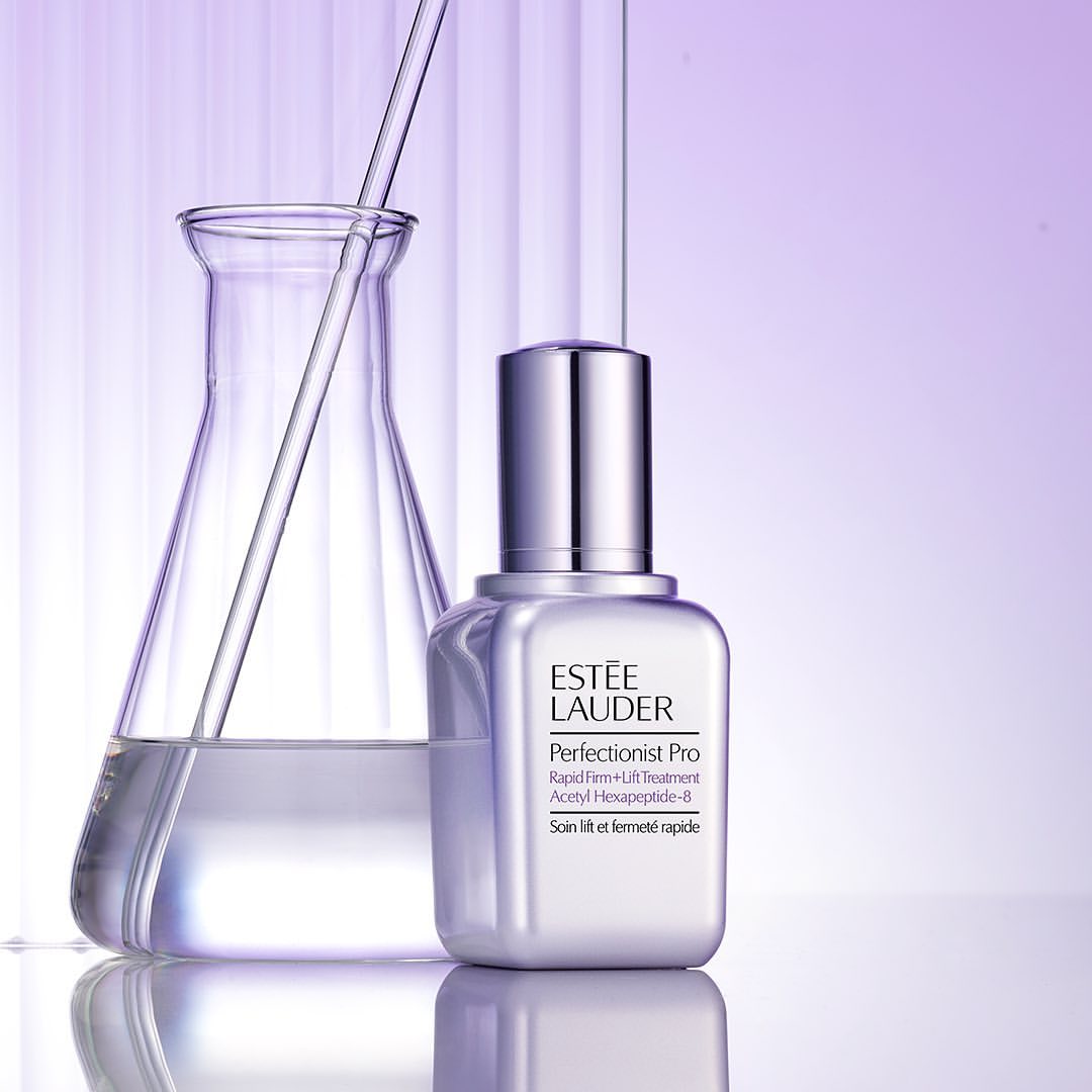 Estée Lauder Perfectionist Pro skincare product shot in a lab-like setting with a beaker and clear liquid against a gradient purple background.