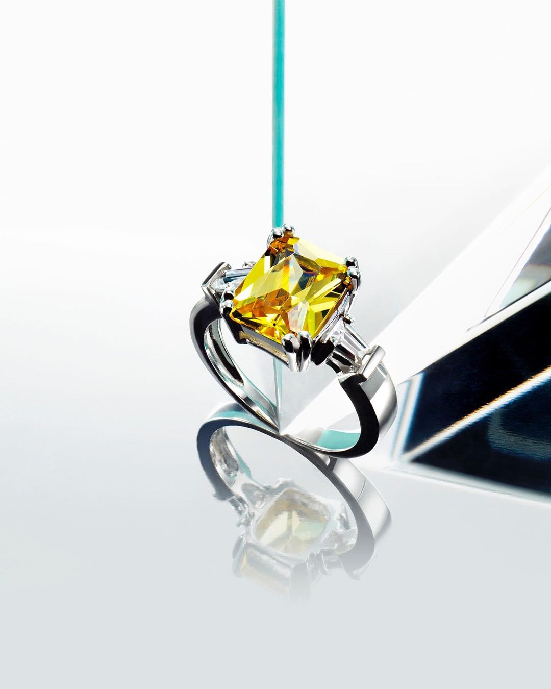 Close-up of a silver ring featuring a square, yellow gemstone and smaller clear gemstones, suspended by a thin blue rod against a white background.