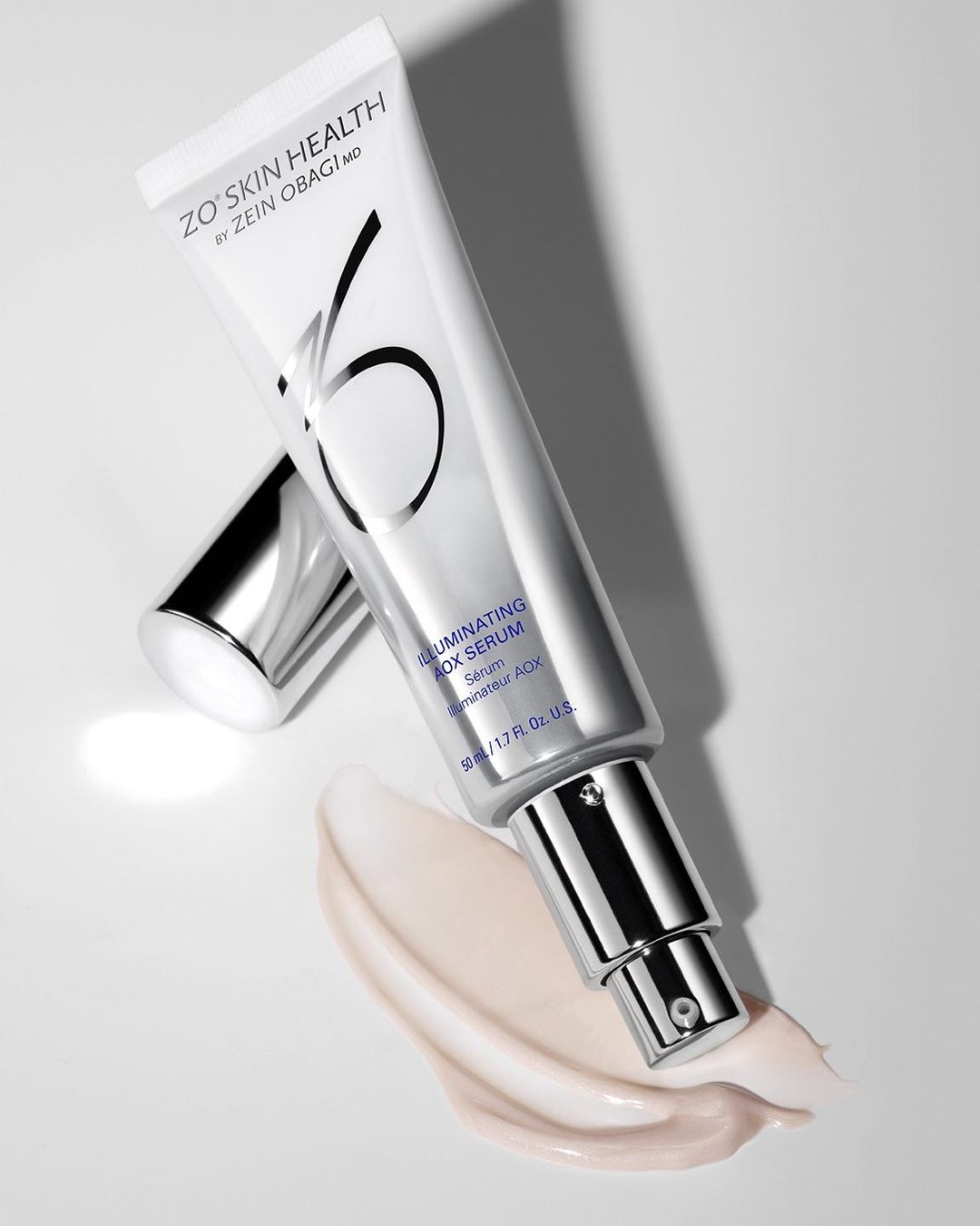 Close-up studio shot of ZO Skin Health Illuminating AOX Serum in a white tube with silver accents, alongside a cream-colored serum swatch.