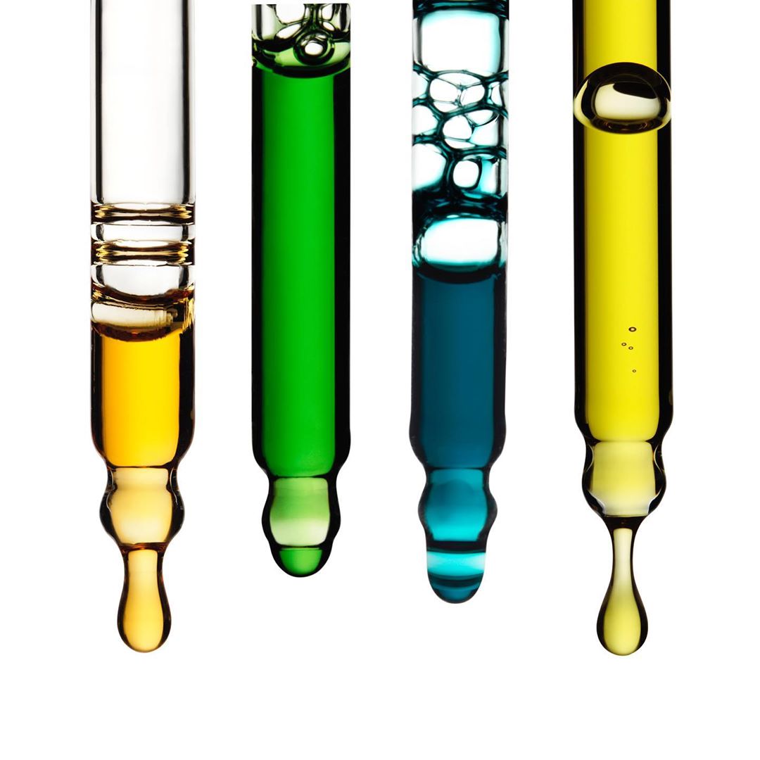 Four glass pipettes with different colored liquids: yellow, green, and blue, dripping from the tip against a stark white backdrop.