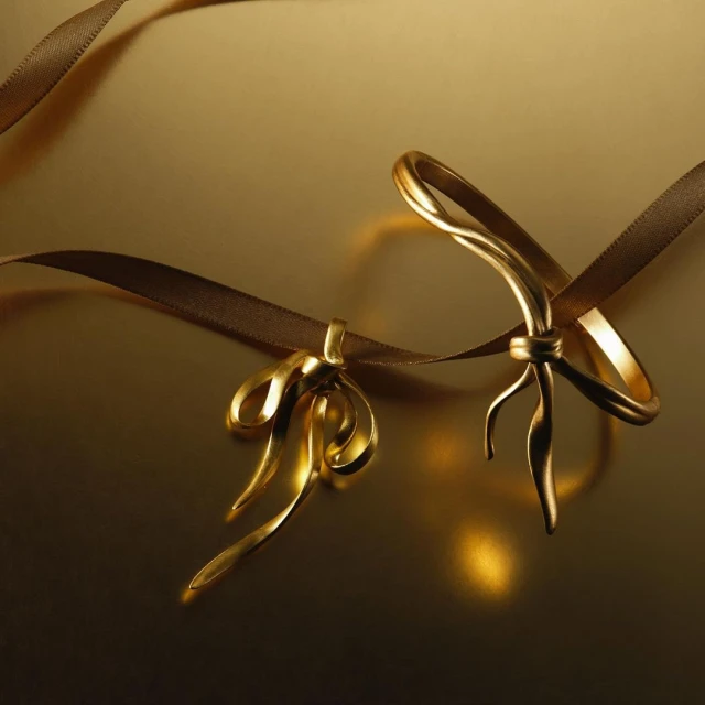 Two golden ribbon bows with a brown ribbon on a reflective golden surface. The scene is lit with soft, warm light and features a shallow depth of field.