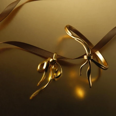 Two golden ribbon bows with a brown ribbon on a reflective golden surface. The scene is lit with soft, warm light and features a shallow depth of field.