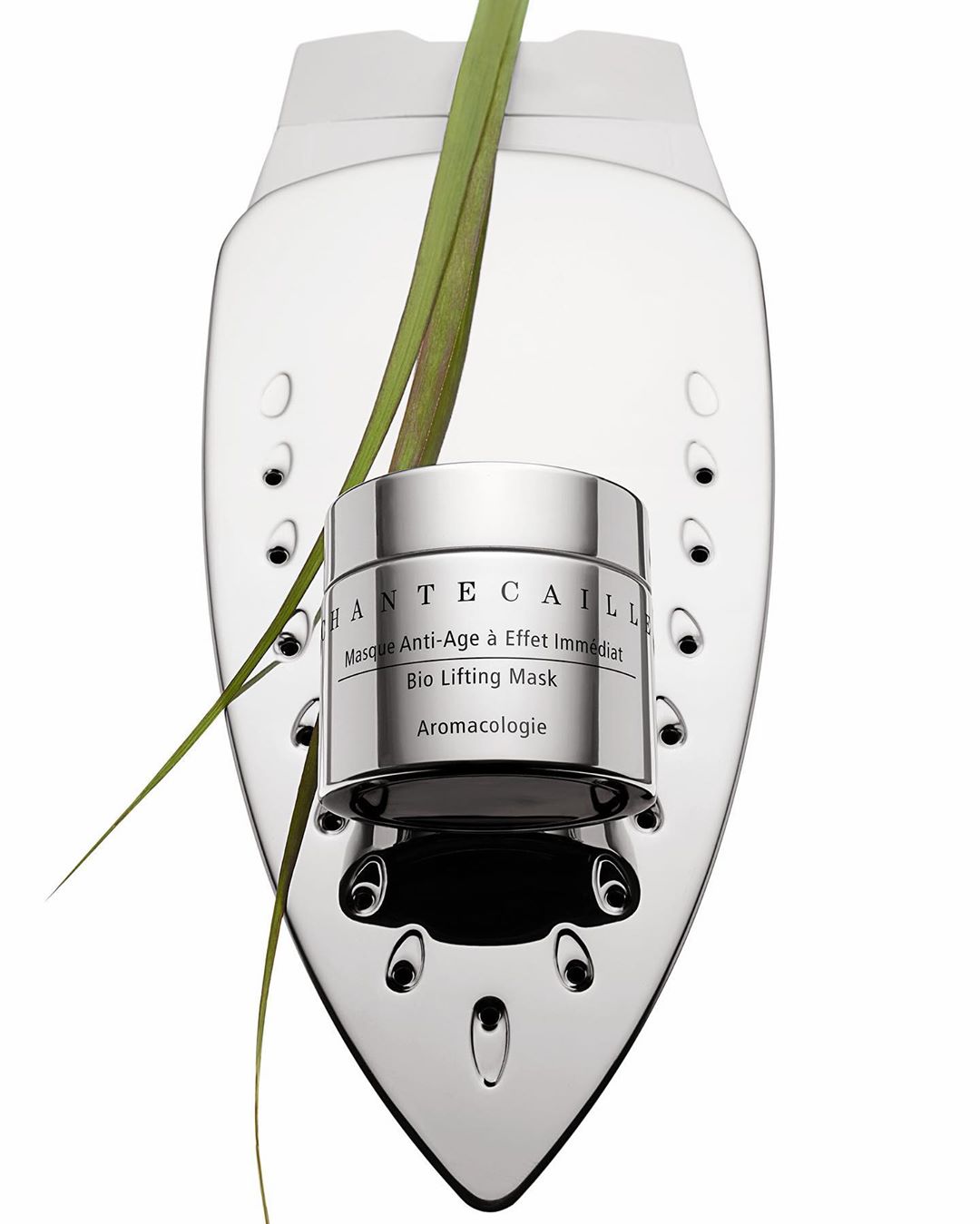 Chantecaille Bio Lifting Mask and green foliage on a chrome stand on a white backdrop, highlighting a luxury beauty product.
