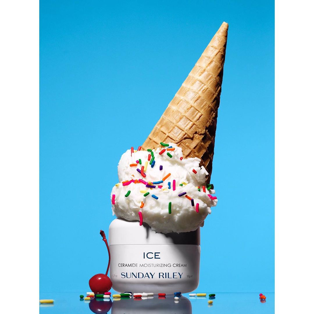 Cosmetic container with ice cream scoop on top, sprinkles, and cherry against a vibrant blue background in a studio setting.