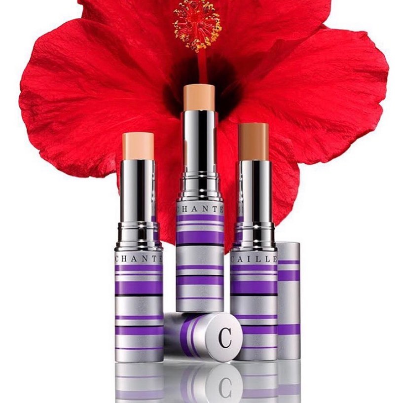 Three Chantecaille foundation sticks in varying shades are displayed with a red hibiscus flower in the background, cosmetics still life.