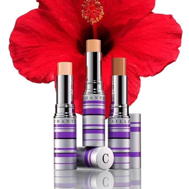 Three Chantecaille foundation sticks in varying shades are displayed with a red hibiscus flower in the background, cosmetics still life.