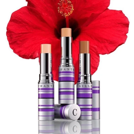 Three Chantecaille foundation sticks in varying shades are displayed with a red hibiscus flower in the background, cosmetics still life.