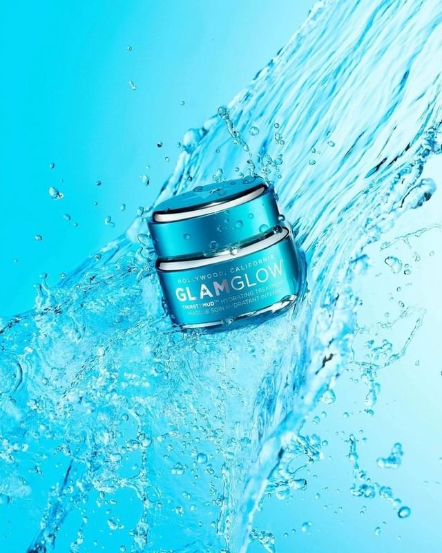 Turquoise GlamGlow skincare containers sit stacked in a splash of clear water against a cool blue background in a beauty product shot.