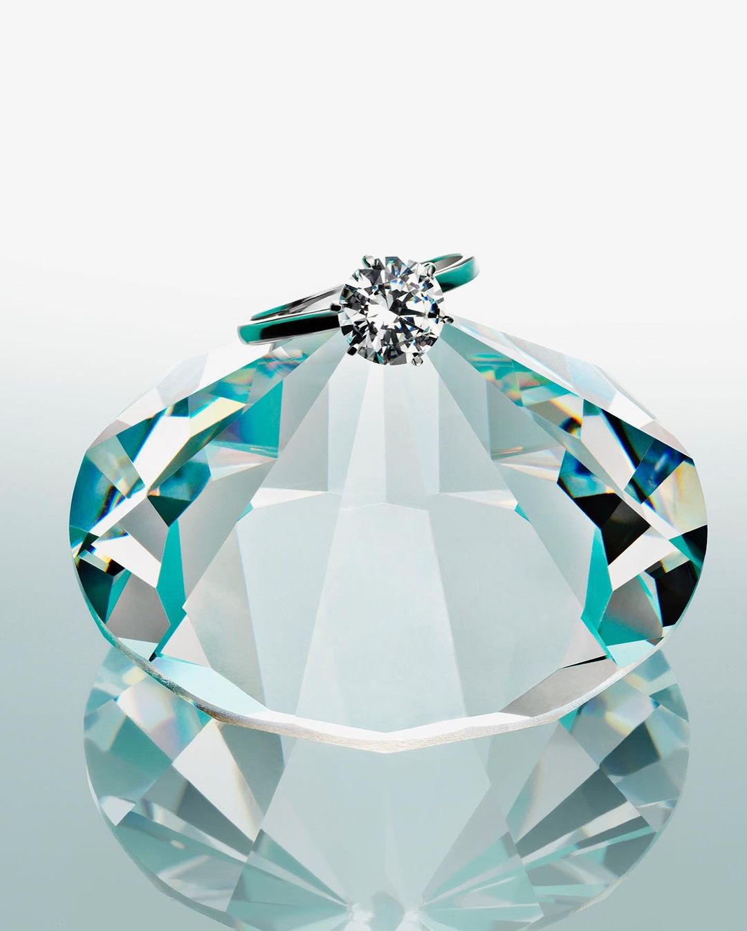 Close-up studio shot of a round-cut diamond solitaire engagement ring on a faceted glass diamond figure against a gradient white background.