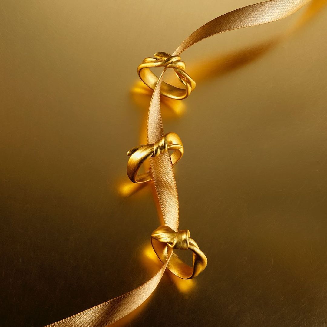 Three gold rings are placed along a light gold ribbon, displayed against a matching gold background in a jewelry studio setting.