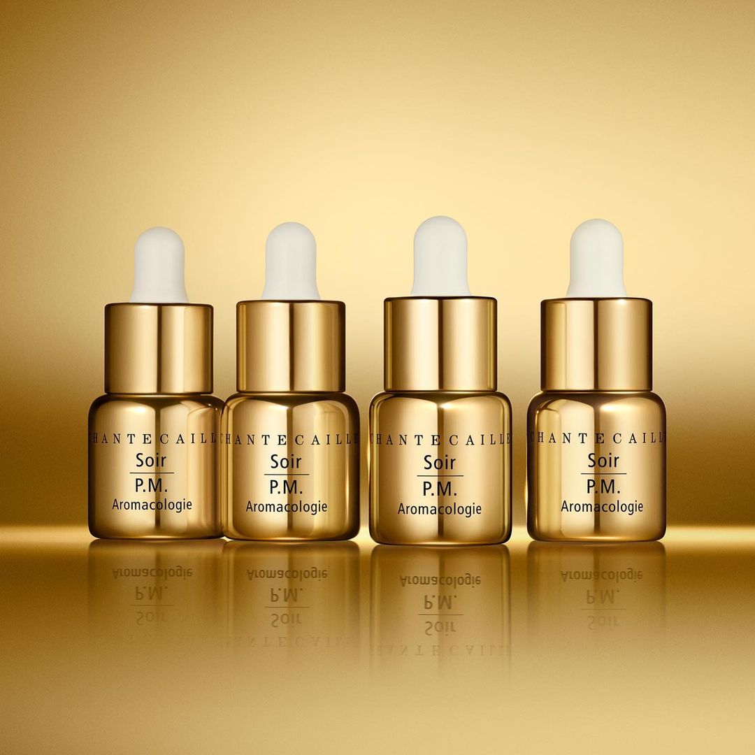 Four gold Chantecaille Soir P.M. Aromacologie bottles with droppers are displayed against a shiny gold background for Beauty advertising.