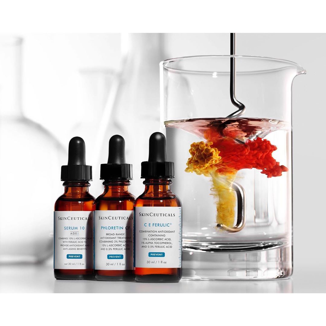 Three SkinCeuticals bottles with droppers stand next to a clear beaker filled with colorful liquid on a white surface in a studio setting.