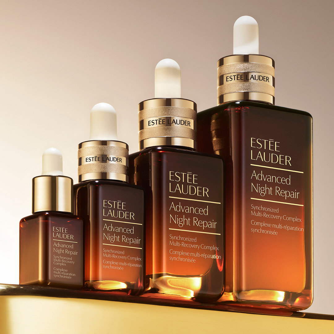 Four amber Estee Lauder Advanced Night Repair serum bottles of different sizes are lined up on a reflective gold surface.