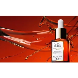 Amber glass bottle of Sunday Riley face oil surrounded by dynamic splashes of golden serum against a rich dark background