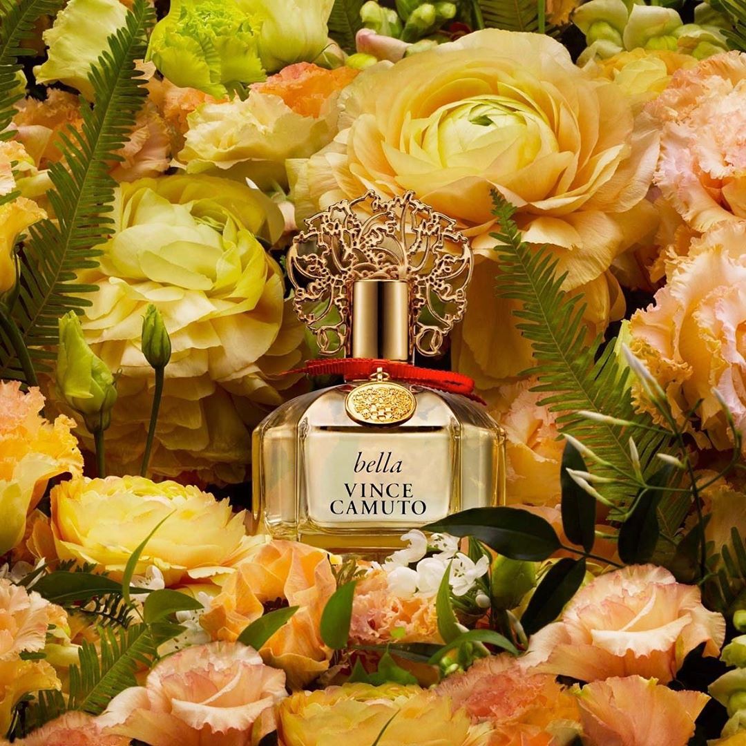 Vince Camuto Bella perfume bottle nestled amidst a dense arrangement of yellow and peach flowers with green fern fronds.