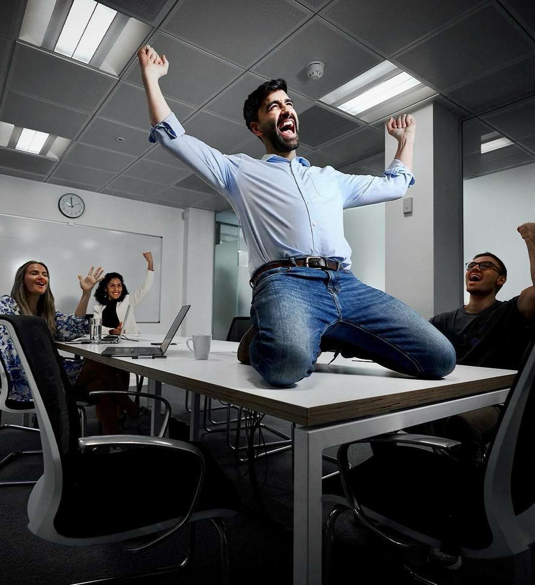 A group of business professionals celebrate in a modern office setting. A man kneels on a table with raised arms in a moment of joy.
