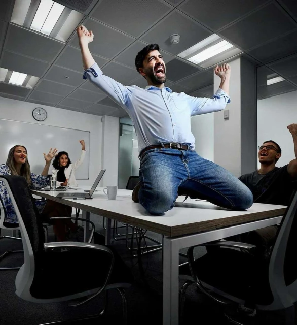 A group of business professionals celebrate in a modern office setting. A man kneels on a table with raised arms in a moment of joy.