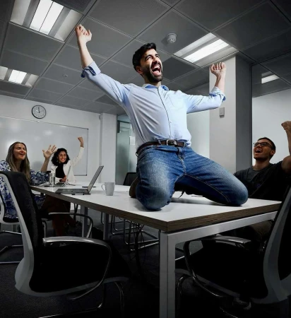 A group of business professionals celebrate in a modern office setting. A man kneels on a table with raised arms in a moment of joy.