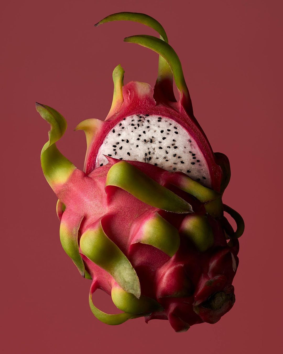 Vibrant, close-up studio shot of a halved dragon fruit, showcasing its white flesh, black seeds, and pink skin against a muted red backdrop.