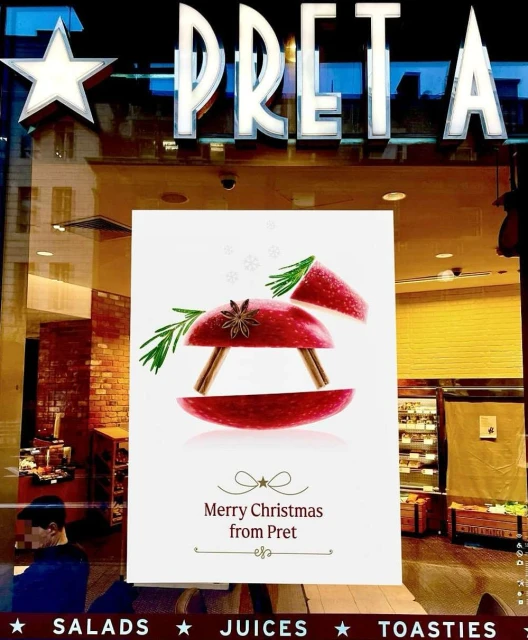 A Christmas poster displayed in a Pret shop window features stylized apple slices with spices, rosemary, and cinnamon sticks.