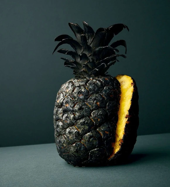 Artistic food shot of a black-painted pineapple with a slice cut out, revealing the bright yellow fruit against a dark background.