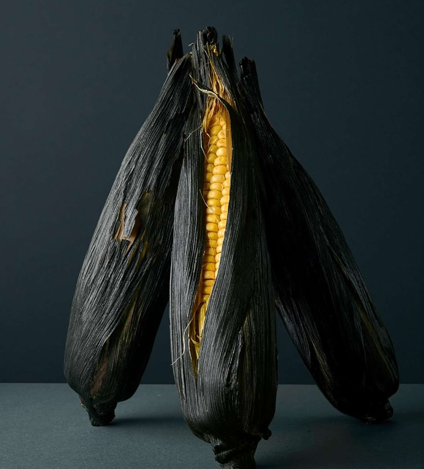 Close-up studio shot of three ears of corn, featuring one partially shucked to reveal yellow kernels, against a dark gray background.