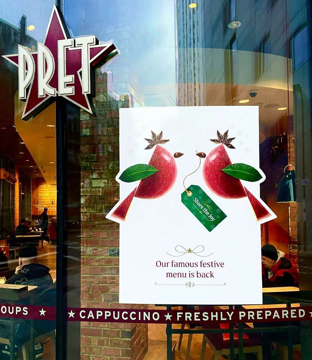 Illustrated holiday advertisement for Pret a Manger on a window, featuring apples, leaves, and star anise, with building reflections.