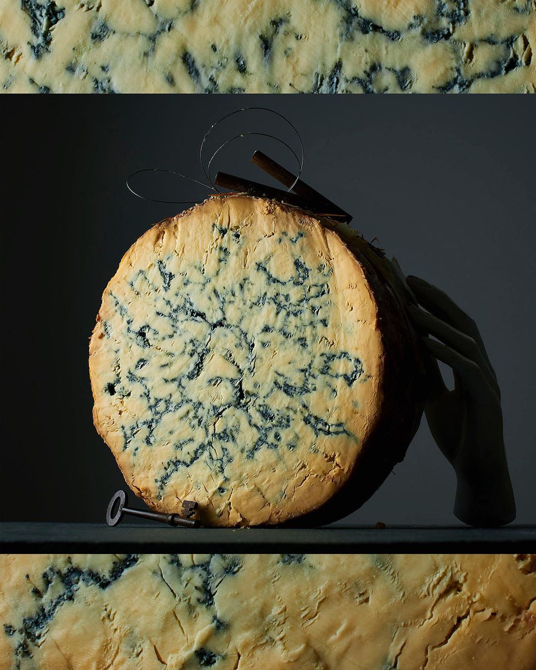 Close-up of a cut wheel of bleu cheese with mold patterns, a metal key, and a decorative metal hairpin against a dark background.