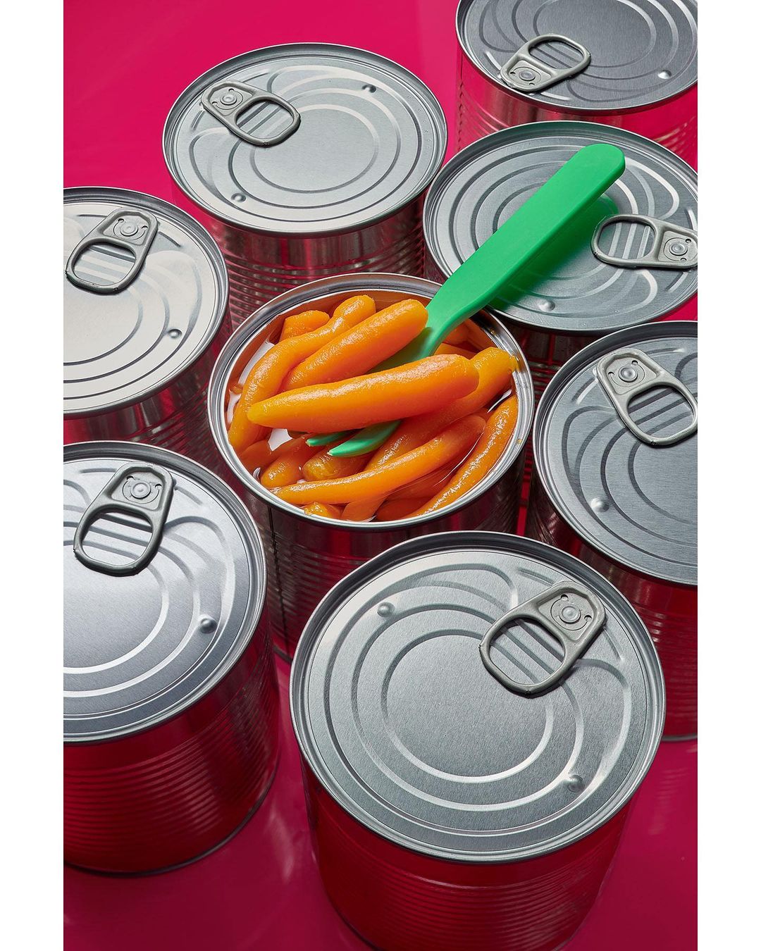 Canned baby carrots surrounded by unopened cans with a green utensil inside, against a bright pink backdrop in a food studio setting.