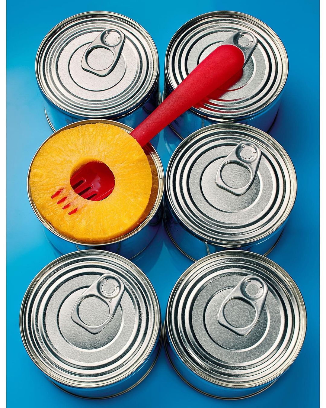 Canned pineapple rings with a red can opener over a light blue background, top-down view, still life food photography.