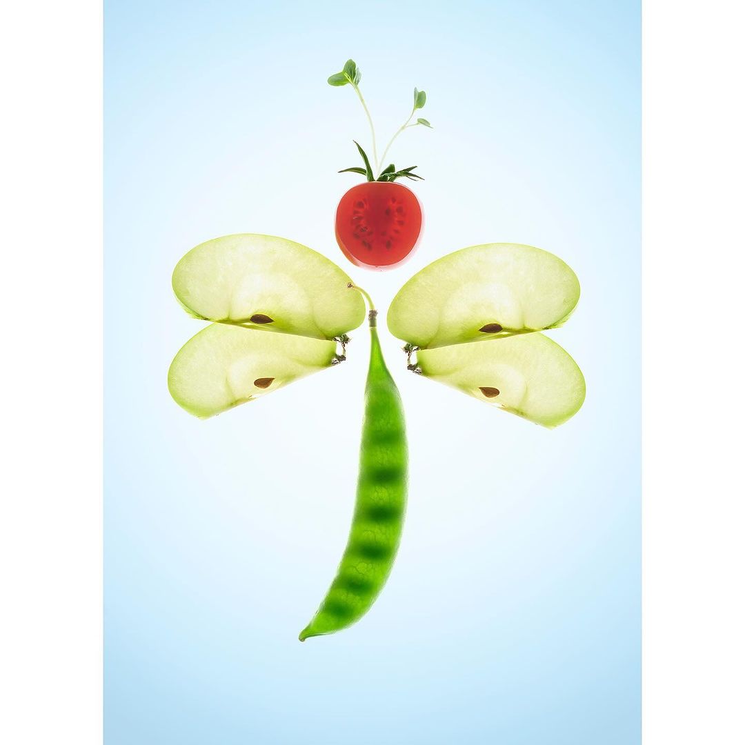 Abstract food art composition with a tomato, pea pod, and sliced apples arranged to resemble a dragonfly on a light blue gradient background.