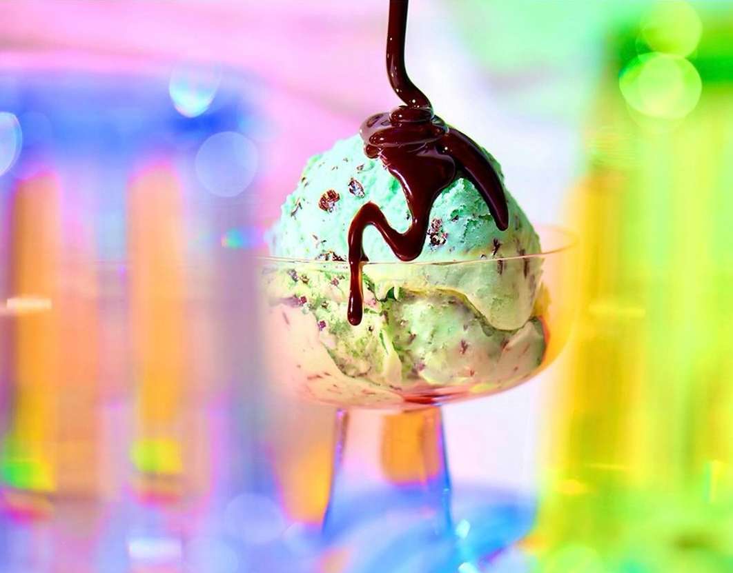 Scoop of mint chocolate chip ice cream in a glass dessert cup, drizzled with chocolate syrup against bokeh background.