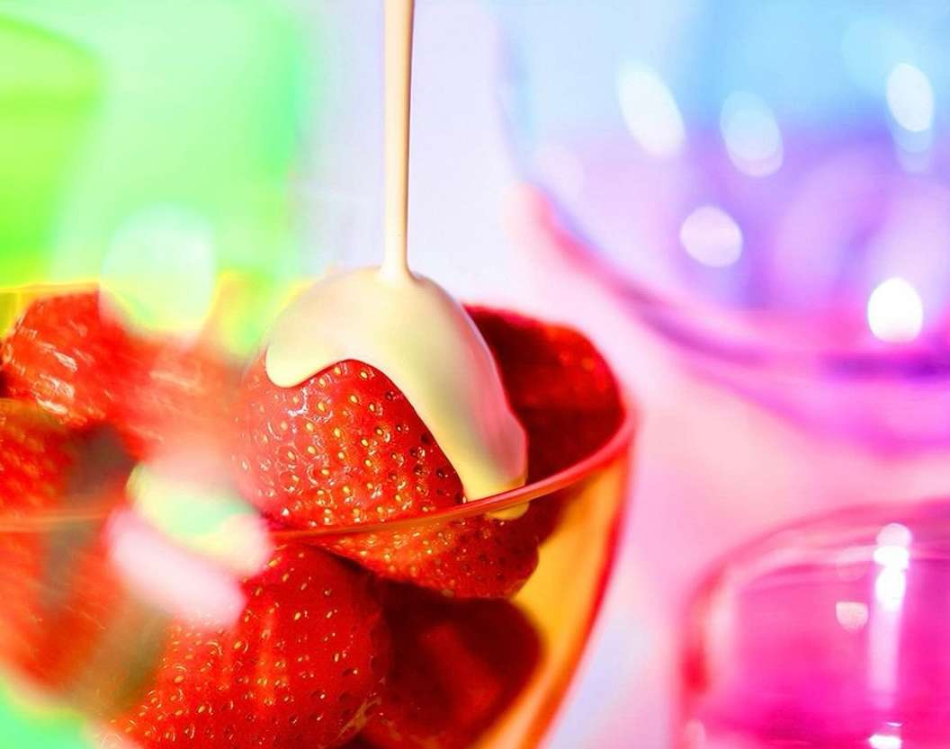 Close-up of fresh strawberries in a glass bowl being drizzled with white cream against a vibrant, multicolored background.