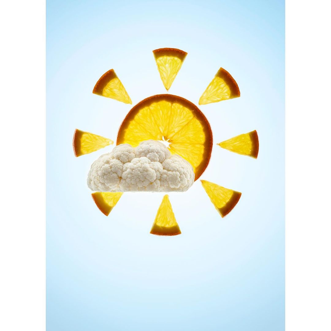 Orange slice sun with cauliflower cloud against blue sky. Food art composition with citrus fruit and vegetable in conceptual weather scene.