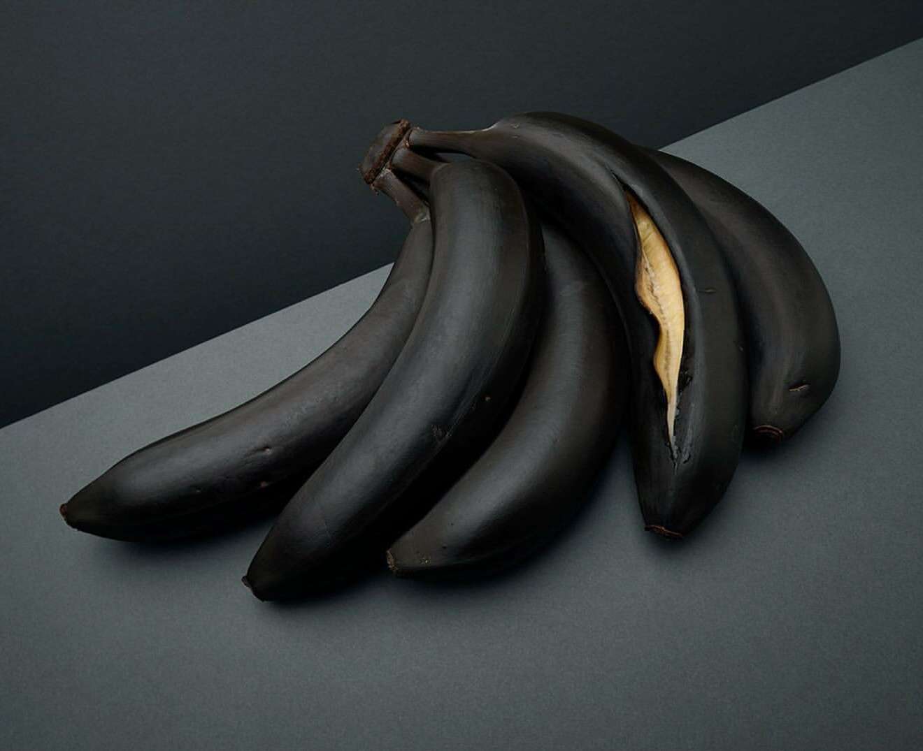 A bunch of dark blue bananas are displayed on a gray two-tone background, with one partly peeled to reveal the yellow flesh.