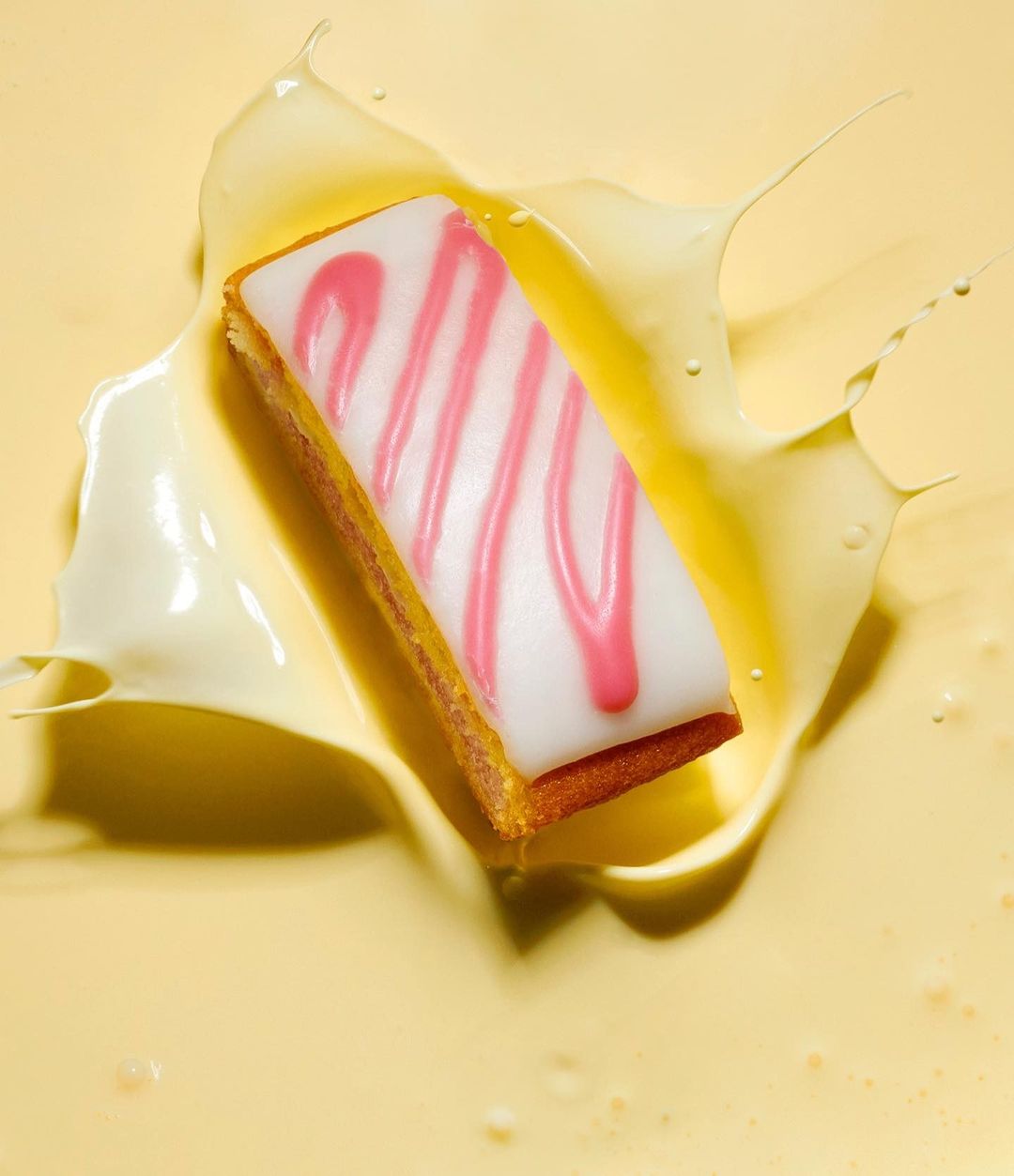 Overhead shot of rectangular sponge cake with white icing and pink drizzle, partially submerged in creamy yellow liquid.