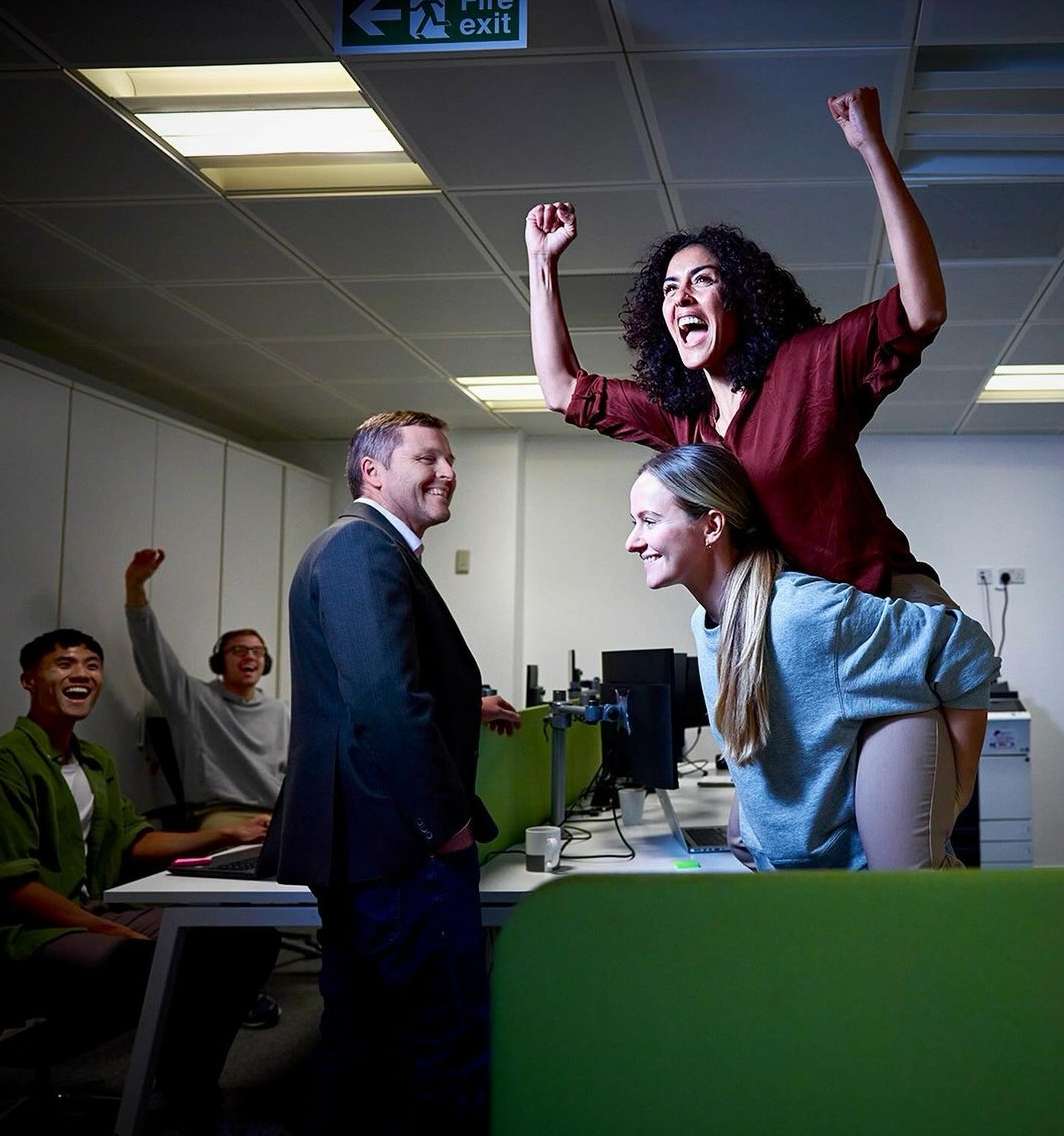 Office celebration: a woman rides piggyback, arms raised in excitement, while colleagues celebrate in an office setting, with a fire exit sign.