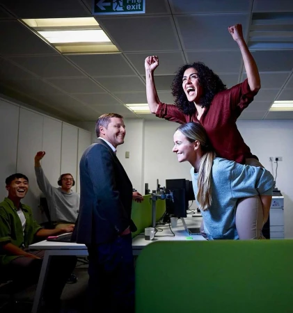 Office celebration: a woman rides piggyback, arms raised in excitement, while colleagues celebrate in an office setting, with a fire exit sign.