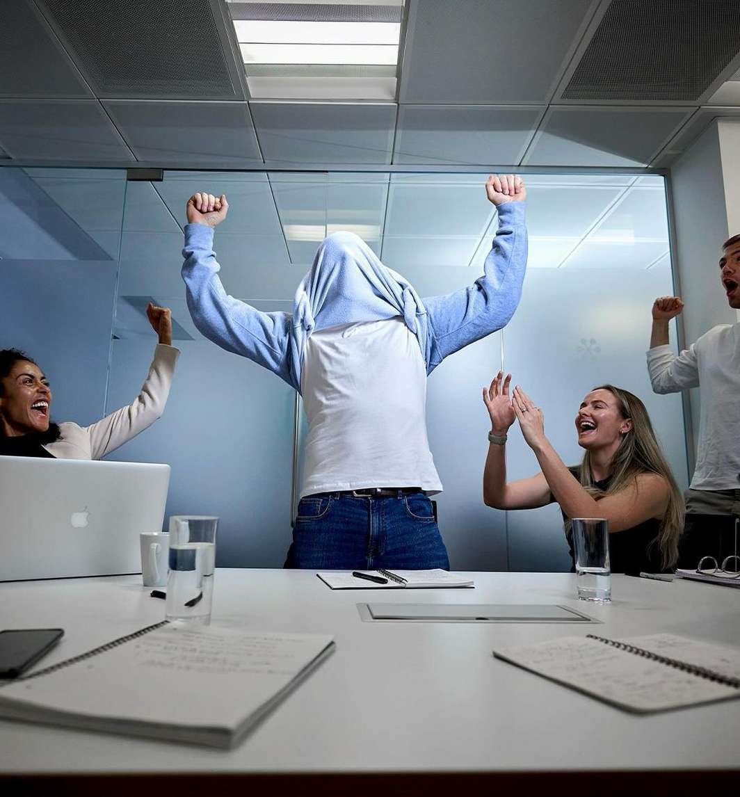 Five smiling office workers celebrate success in a brightly lit conference room around a white table with laptops, pens, and notepads.