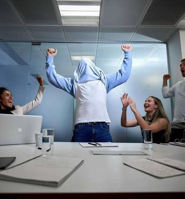 Five smiling office workers celebrate success in a brightly lit conference room around a white table with laptops, pens, and notepads.