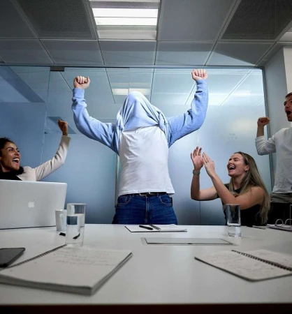 Five smiling office workers celebrate success in a brightly lit conference room around a white table with laptops, pens, and notepads.