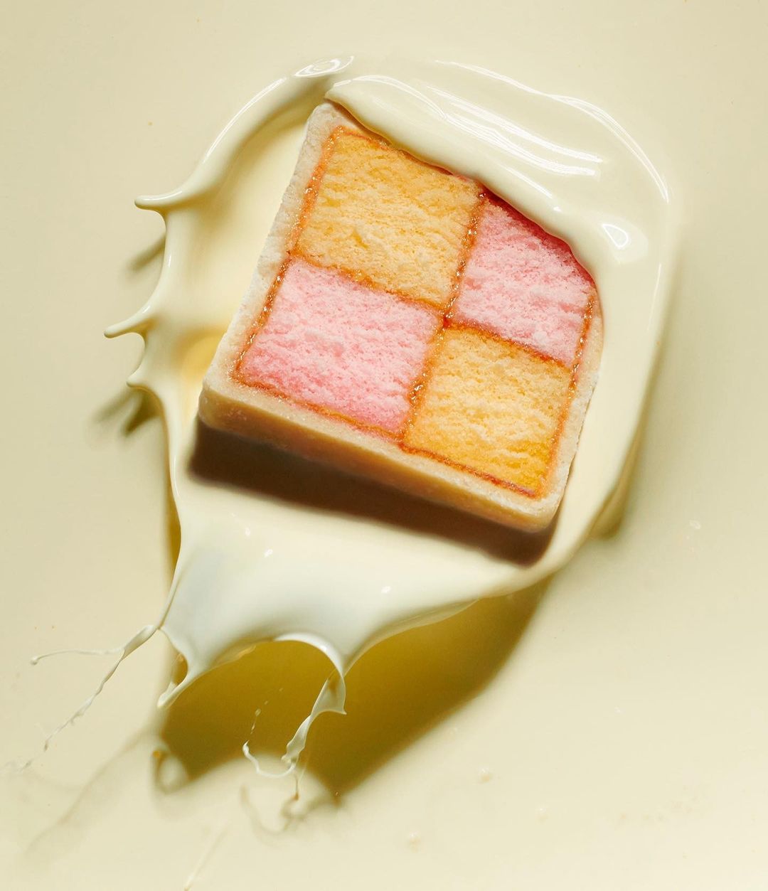 Close-up of frosted sponge cake with pink and yellow checkerboard pattern, partially submerged in white liquid, macro studio shot.