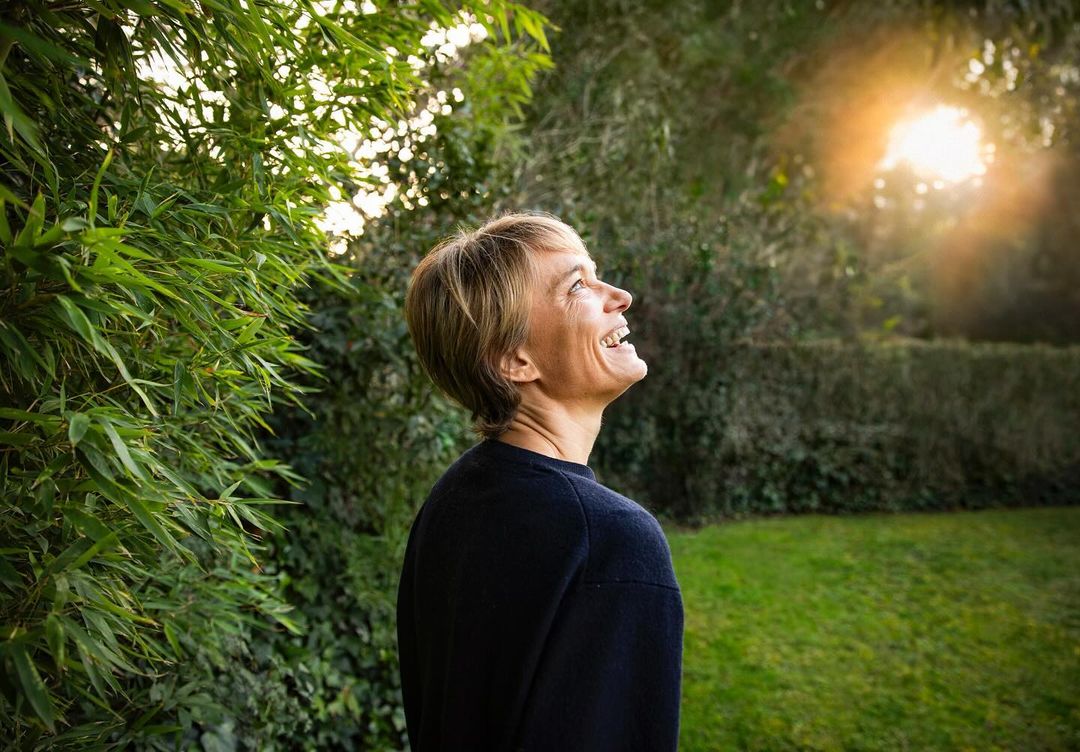 Adult woman smiles looking up towards the sun in outdoor setting surrounded by greenery and trees with shallow depth of field.