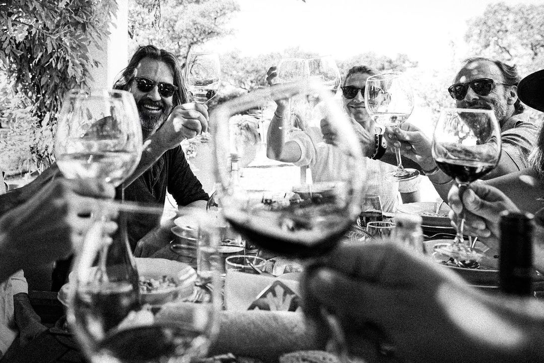 Black and white photo of group raising wine glasses in a toast, focusing on smiling faces and the gathering in a sunlit outdoor setting.