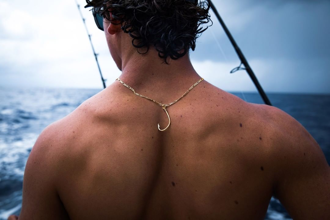 Bare-chested man on a boat with a hook necklace, fishing rods, and ocean backdrop. Sport fishing lifestyle, open water, and nautical theme.