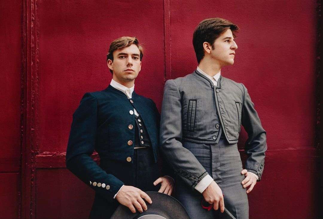 Two adult males in matching formalwear pose in front of a red wall, one looking forward, the other glancing to the side.