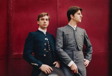 Two young men in traditional tailored jackets standing against a deep red wall, photographed by Richard Schultz.