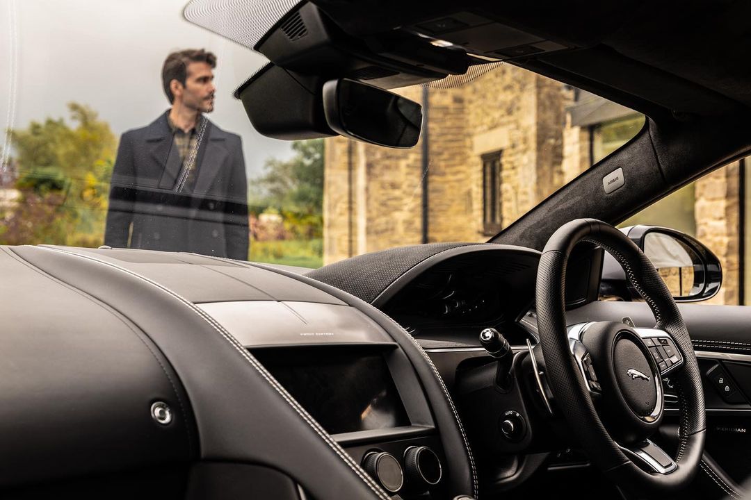 Car interior showcases sleek black design with leather textures and dashboard details; building visible with blurred person through windshield.