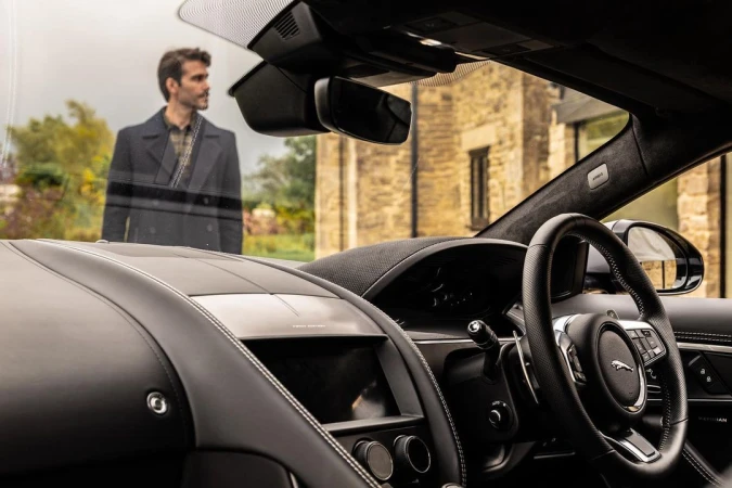 Car interior showcases sleek black design with leather textures and dashboard details; building visible with blurred person through windshield.